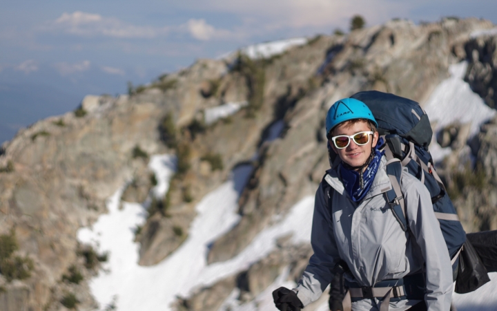 a young person wearing a helmet and a backpacking smiles at the camera. There is a snowy and rocky landscape in the background.